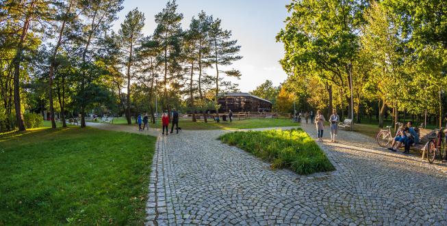 Zdjęcie wykonane w wiosenny dzień w Parku Zdrojowym. Ścieżką spacerową idą turyści, na ławce po prawej stronie siedzą rowerzyści. W głębi widać wysokie zielone drzewa oraz tężnie solankową. 