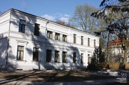 Dwukondygnacyjny budynek ze zdobioną jasną elewacją, z prawej strony drzewa