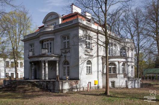 Na środku willowy budynek piętrowy, od frontu widoczne wejście z zaokrąglonym daszkiem wspartym na kolumnach, do którego prowadzą schody, dach z mansardowymi oknami, pokryty dachówką w kolorze czerwonym, z lewej strony widoczny fragment innego budynku