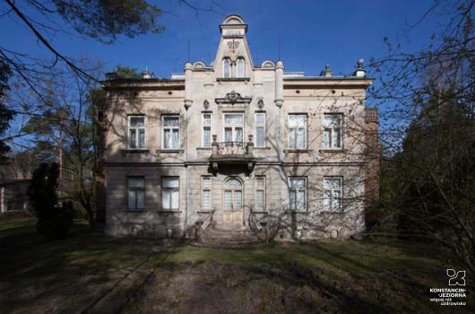 Pośrodku widoczny piętrowy okazały budynek willowy, z ozdobnym wejściem i zdobieniami na froncie,, balkonem nad wejściem i fryzem z napisem JULIA, elewacja jasna, do willi prowadzi droga, wokół drzewa