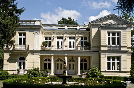 Pietrowy budynek willowy w stylu neoklasycystycznym, nad wejściem taras podparty kolumnami, dach z częsciową kolumnadą, przed budynkiem fontanna z rzeźbą