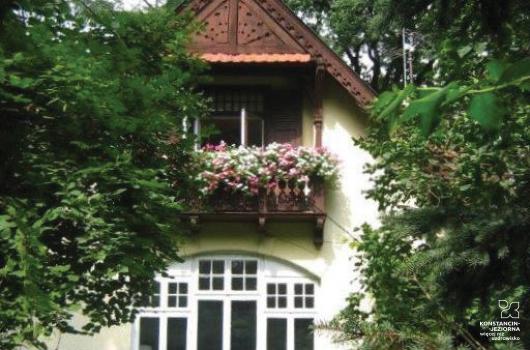 Widoczny fragment frontowej części budynku ze spadzistym dachem i balkonem nad dużym oszklonym wejściem, boki zasłaniają drzewa