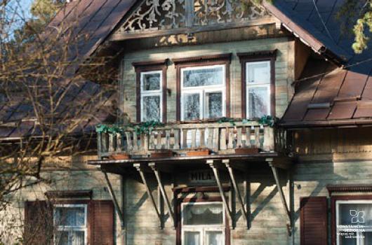 Widoczny fragment frontu drewnianego jednopoziomowego budynku, z dachem spadzistym, krytym blachą, na środku dachu nadbudówka z balkonem nad wejściem, z bogatym zdobieniem snycerskim