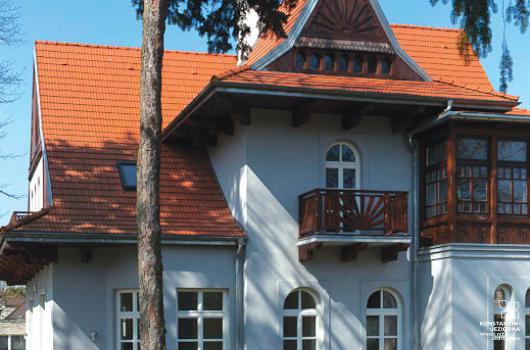 Duży fragment budynku willowego, parterowego z dużym stromym dachem i poddaszem mieszkalnym, na dachu czerwone dachówki, na froncie nadbudówka wysoka, front jasny, po prawej stronie budynku dobudowana drewniana weranda, przed budynkiem wysokie drzewa