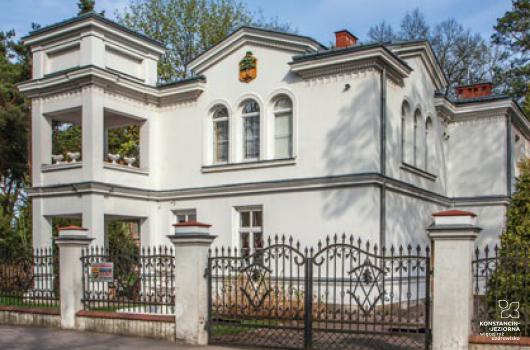 Na pierwszym planie fragment ulicy i ogrodzenie z bramą, za którym, jest biały budynek, piętrowy, z lewej strony budynku dostawiona niby wieża, czworoboczna, ażurowa, budynek z fryzami bogato zdobionymi