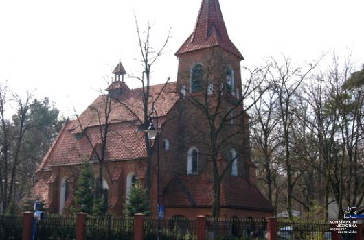 Budynek kościoła z czerwonej cegły i z czerwonym dachem, widziany z przedniego narożnika, z przodu wejście do budynku z nadbudowaną wieżą zakończoną strzelistą kopułą