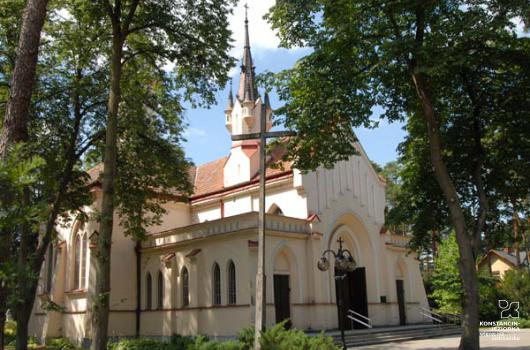 Budynek kościoła, ze strzelistą zdobioną wieżą, wejście i okna z ostrymi zwieńczeniami nawiązującymi do gotyku, wokół kościoła drzewa