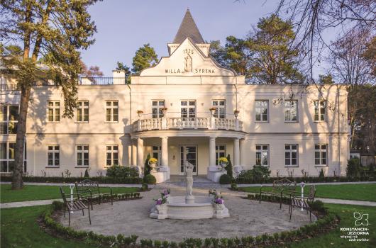 Okazały budynek willowy, dwukondygnacyjny, nad drzwiami wejściowymi balkon z kolumnadą, przed wejściem widoczna fontanna