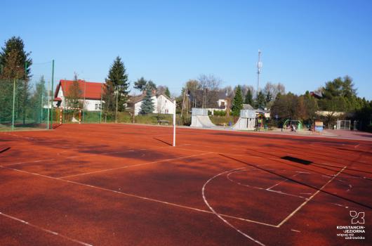 Boisko z czerwoną nawierzchnią, z wyrysowanymi liniami, w tle widoczne zabudowania