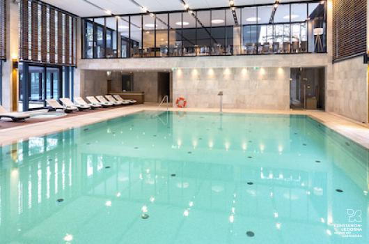 Duży basen w jasnej hali, ściany po lewej i prawej szklane, po prawej stronie stoją leżaki i widoczne jest jacuzzi