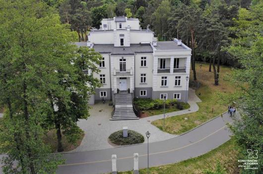 Duży pi,etrowy obiekt willowy widziany z lotu ptaka, od frontu z góry, wokół drzewa, przed budynkiem i wzdłużfrontu sieżki