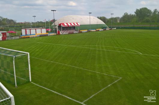 Duże boisko do piłki nożnej, z wyrysowanymi liniami, po lewej stronie widoczne trybuny i duży namiot