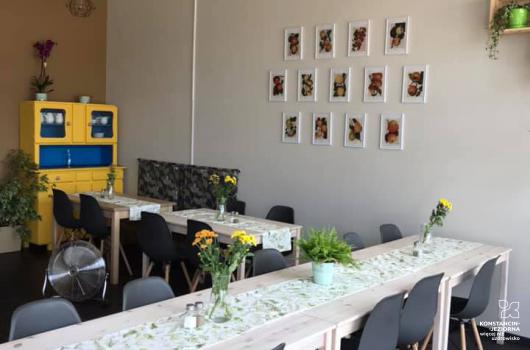 Wnetrze małego lokalu, stoliki poustawiane w literę L, z dostawionymi krzesełkami, na stołąch obrusy i kwiatki w wazonach, na ścianie po prawej wiszą obrazki w ramkach, w rogu stoi stary kredens w klorze żółtym