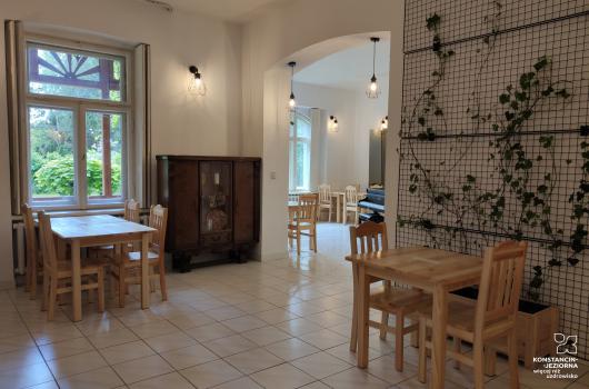Wnetrze sali restauracyjnej, drewniane stoliki i krzesełka, podłoga terrakota jasna, z lewej strony duże okno, po prawej ściana z siatką i bluszczem, pośrodku łukowate przejście do innego pomieszczenia z widocznym fortepianem