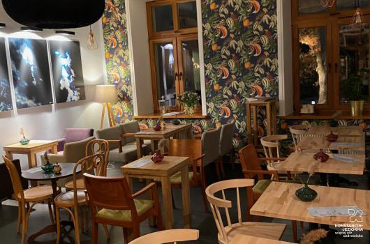 Wnętrze kawiarni z drewniamymi stolikami i krzesłami, ściana z florystyczną tapetą, po prawej widoczne okna, po lewej na ścianie duże fotografie, spod sufitu wiszą lampy