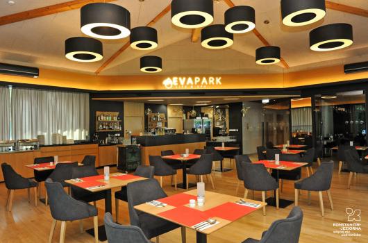Wnetrze nowoczesnej restauracji, pośrodku nakryte stoliki z fotelikami, naśrodku i po lewej stronie widoczny bar z zastawą, po prawej przeszkolne dzrwi wejściowe, spod sufitu zwisają koliest lampy, pośrodku nad barem neon z nazwalokalu