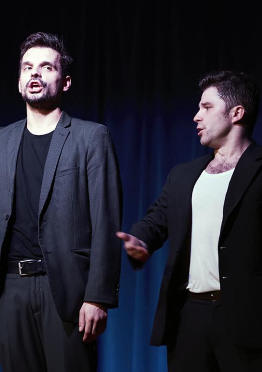 Scena teatralna. Dwóch mężczyzn na scenie. Stoją obok siebie i coś mówią. W tle widać kurtyny sceniczne.