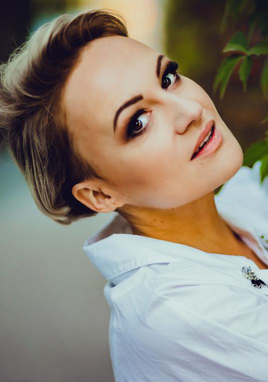 Młoda kobieta, długie włosy związane w kok. Ubrana jest w białą koszulę. Stoi przy drzewie.