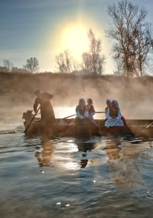 Drewniana stara łódź płynąca na rzece. Na niej znajduje się 6 osób – 4 kobiety ubrane w stroje ludowe i dwóch mężczyzn trzymających wiosło. W tle widać wysokie drzewa. 