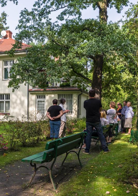 Zabytkowa willa – duża ilość okien, spadzisty czerwony dach, jasna elewacja. Przed budynkiem stoi grupa osób. 