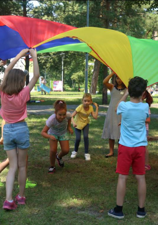 Park. Grupa dzieci w wieku szkolnym stoi w okręgu, w rękach trzymają dużą kolorową chustę. W tle widać wysokie drzewa.  