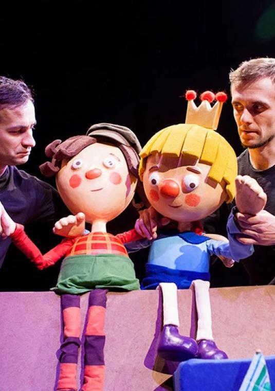 Dwóch młodych mężczyzn - aktorów stoi na scenie. Ubrani są na czarno, pomiędzy nimi siedzą dwie duże lalki teatralne.