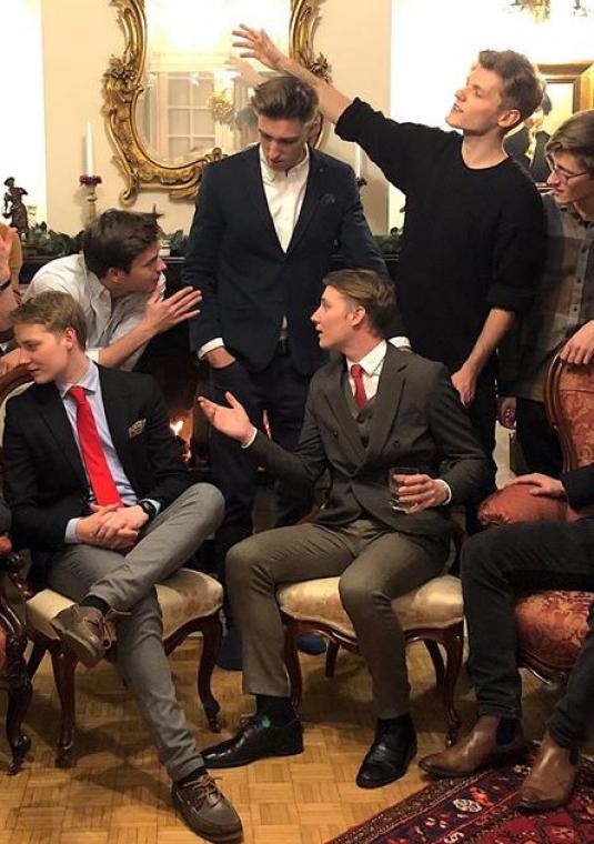 Pokój. Grupa jedenastu młodych mężczyzn rozmawia ze sobą. Czterech mężczyzn siedzi na krzesłach, pozostali stoją za nimi. Wszyscy ubrani są elegancko.