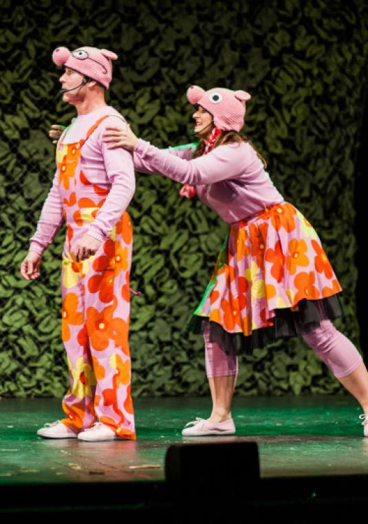 Plakat promujący premierę spektaklu. Trzy osoby stoją obok siebie na scenie. Mężczyzna i dwie kobiety. Ubrani są w pomarańczowo-różowe stroje. Na głowach mają różowe czapki przedstawiające głowę świnki.