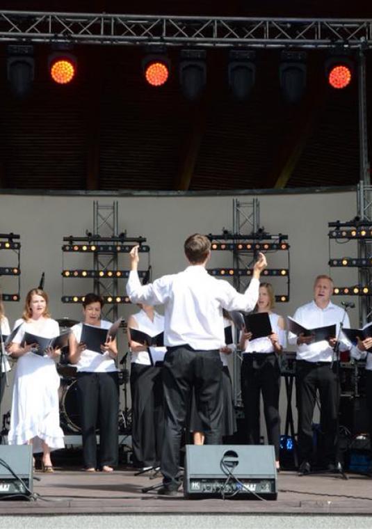 Kilkanaście śpiewających osób stoi na scenie, w półkolu, każdy odświętnie ubrany, każdy trzyma przed sobą otwarty zeszyt z nutami, przed nimi młody mężczyzna z podniesionymi rękami, dyrygujący zespołem, nad nimi widoczne oświetlenie sceniczne