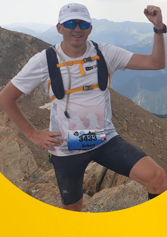 Mężczyzna w sportowym stroju i z umerem startowym stoi na tle szczytów górskich, podnosi w górę lewą rękę z zaciśniętą pięścią, na twarzy ma ciemne okulary, na głowie czapkę z daszkiem