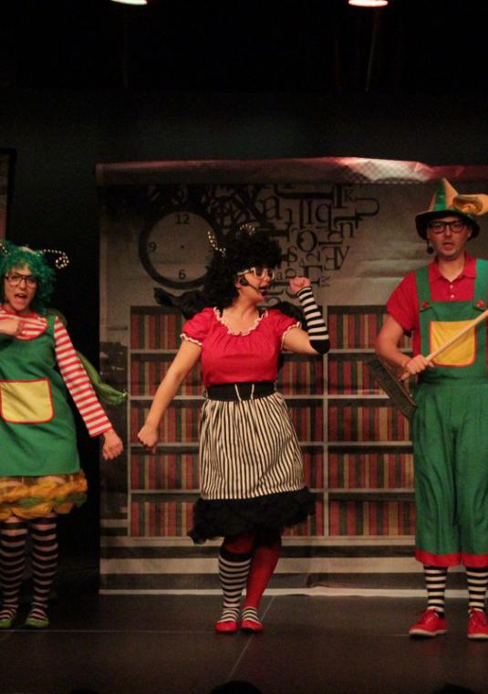 Zdjęcie z opisywanego przedstawienia, trzy żartobliwie i krzykliwie ubrane postaci na scenie teatralnej