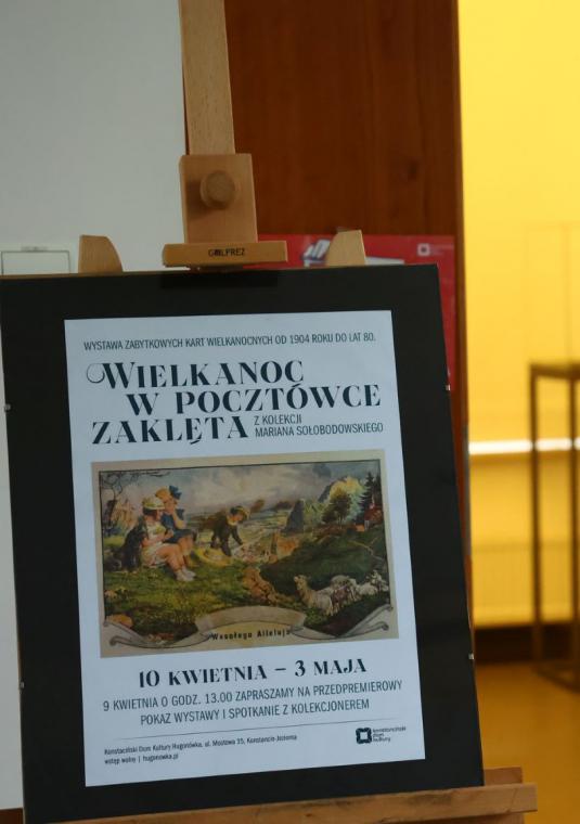 Na stojaku znajduje się plakat promujący wystawę, w tle widać dwie osoby oglądające wystawę. 