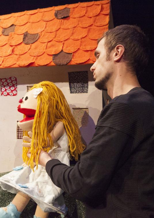 Bajkowa scenografia teatralna, na scenie aktor który w rękach trzyma duże lalki szmaciane.