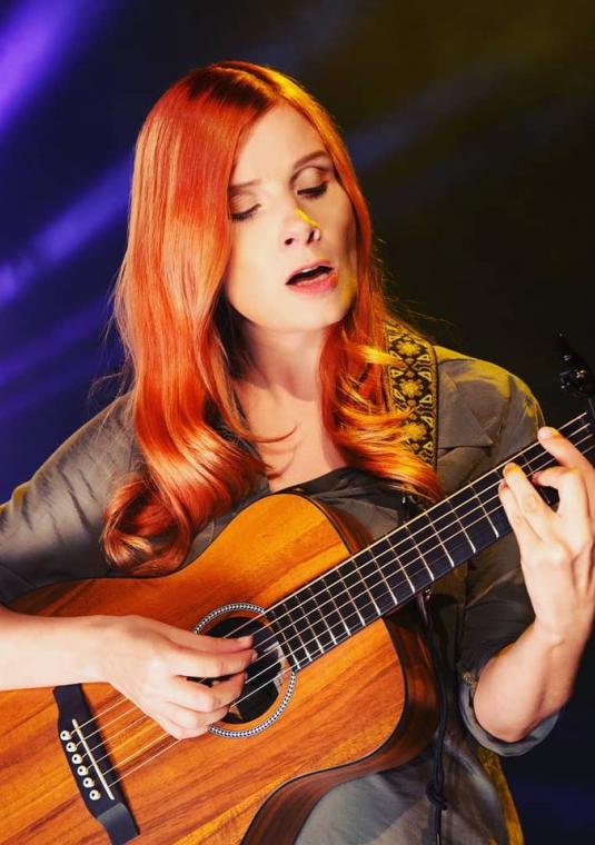 Młoda kobieta, z długimi rudymi włosami gra na gitarze.