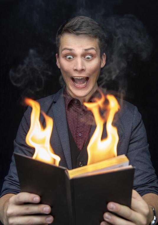 Młody mężczyzna, który z przerażeniem patrzy na płonącą otwartą książkę, którą trzyma w dłoniach.