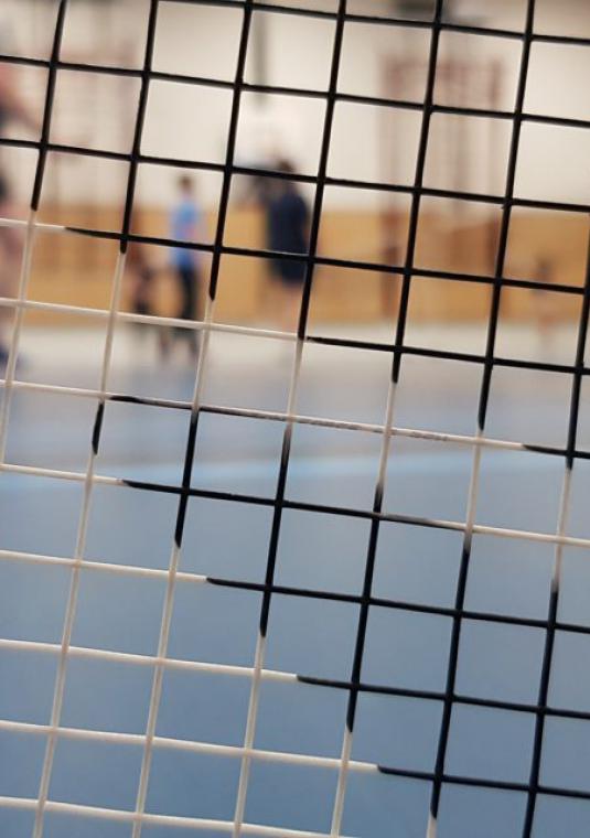 Na zbliżeniu siatka rakietki do badmintona. Przez nią widać sylwetki trzech osób na sali gimnastycznej.