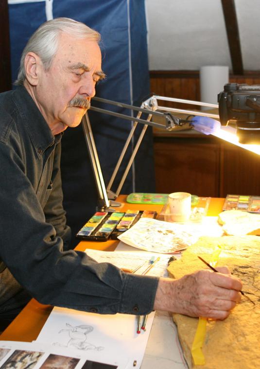 Starszy mężczyzna siedzi za stołem oświetlonym lampami. W dłoni trzyma pędzelek. Na stole leżą farby i kartki.