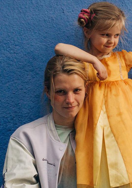 Młoda kobieta i mała dziewczynka w pomarańczowej sukience stoją na tle niebieskiej ściany. 