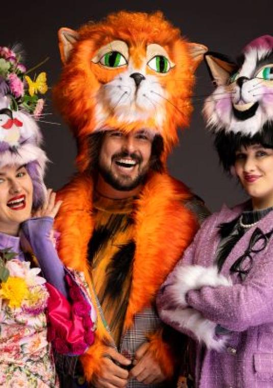 Mężczyzna i po jego bokach dwie młode kobiety. Wszyscy przebrania za koty: w kolorowych kostiumach teatralnych (m.in. maski).