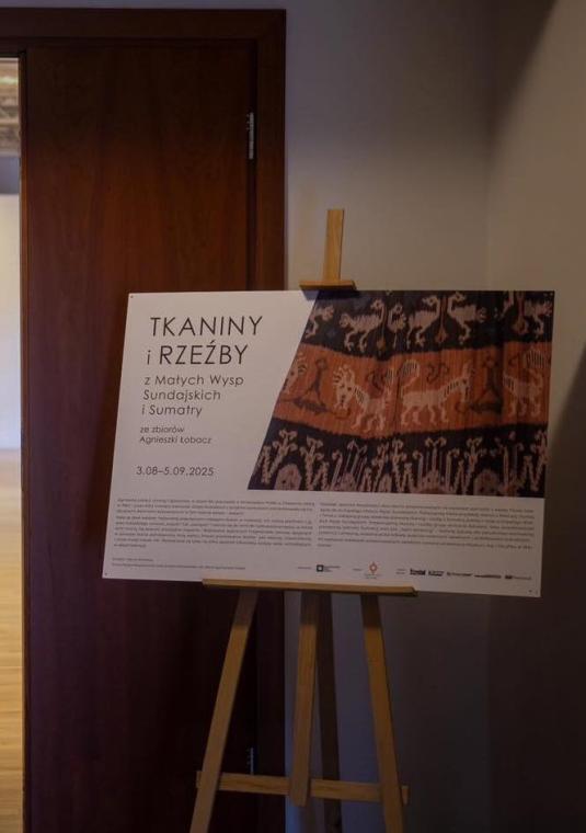 plakat Tkaniny i rzeźby ustawiony na sztaludze obok drzwi prowadzących na wystawę