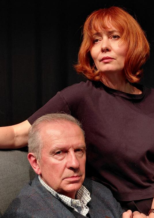 Zdjęcie kobiety i mężczyzny siedzących na szarym fotelu. Czarne tło.