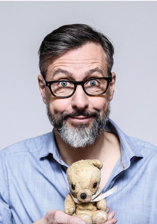 zdjęcie portretowe Szymona Majewskiego, patrzy na nas z figlarnym uśmieszkiem, w rękach trzyma starego, białego pluszowego misia