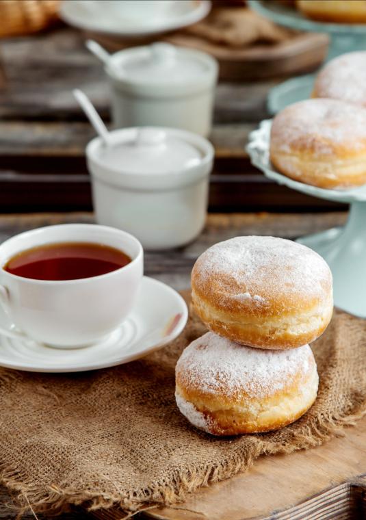 zdjęcie apetycznych pączków obsypanych cukrem pudrem i porcelanowa filiżanka z herbatą, w tle w nieostrości, platera z pączkami i cukiernica