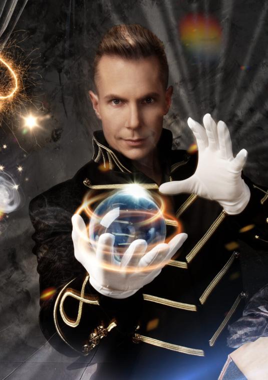 Magiczna grafika przedstawiająca iluzjonistę w czarnym, galowym stroju ze złotymi lamówkami i białych rękawiczkach. Artysta trzyma w dłoniach lewitującą, świecącą kulę. Wokół unosi się dym, karty do gry, płonące symbole karciane (pik i karo), białe pióro, czerwona róża oraz stara księga z czerwonym piórem.