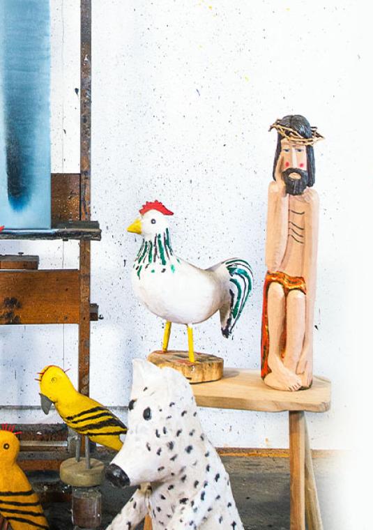 Plakat wystawy „Stanisław Baj i Przyjaciele” w Muzeum Wycinanki. Na zdjęciu widoczne są kolorowe rzeźby ludowe (ptaki, postać Chrystusa) oraz obraz na sztaludze w pracowni artystycznej.