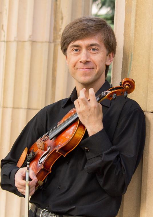 Mężczyzna o jasnej karnacji w czarnej koszuli, trzymający skrzypce i smyczek, opierający się o klasyczną kolumnę.