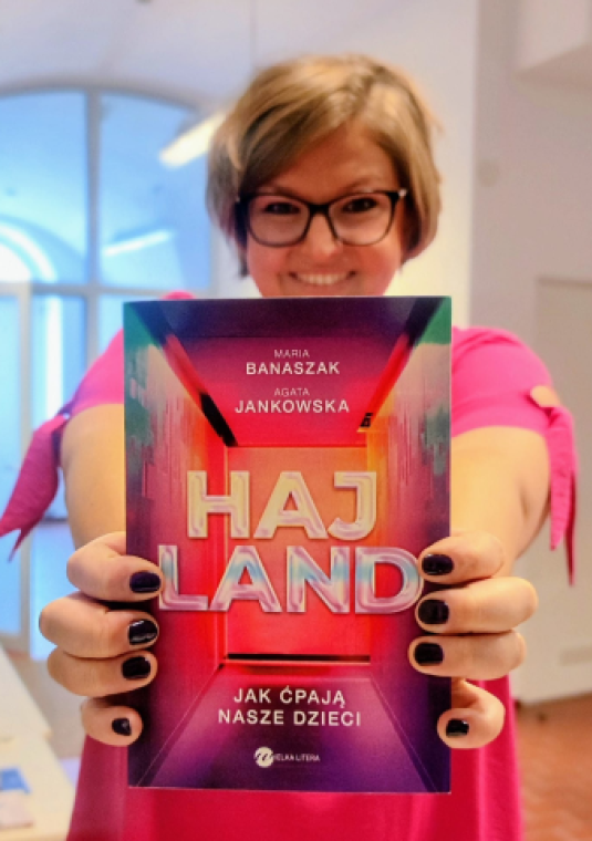 Zdjęcie uśmiechniętej kobiety, która trzyma przed sobą książkę, na okładce napis Haj Land