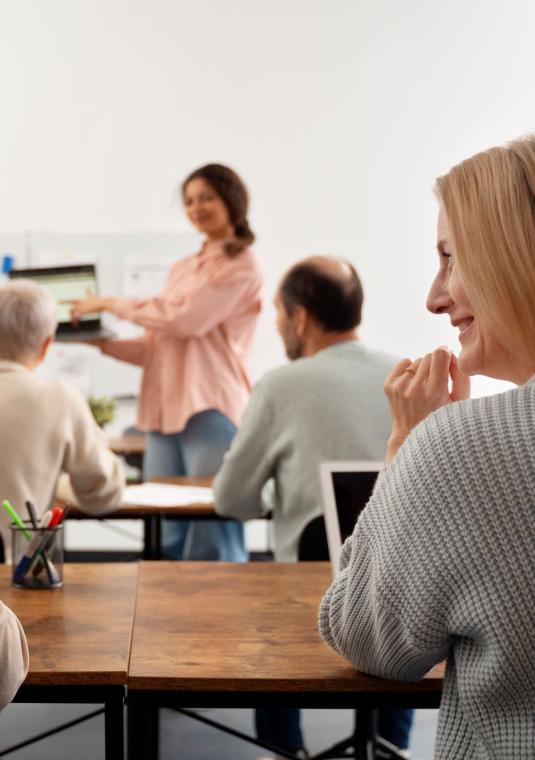 dwoje ludzi siedzi przy stole i patrzą na siebie, w tle widać nauczycielkę trzymającą laptopa