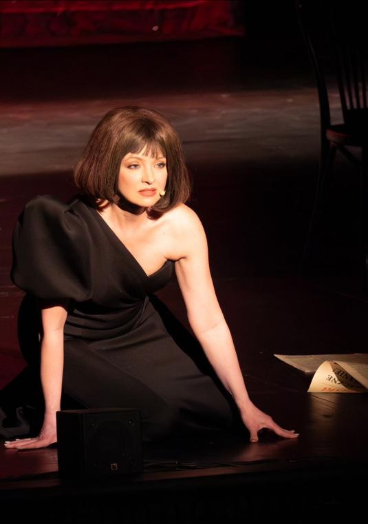 Zdjęcie ze spektaklu teatralnego przedstawiające Barbarę Kurdej-Szatan na scenie, wcielającą się w rolę Kaliny Jędrusik. Aktorka siedzi na deskach sceny w blasku punktowego reflektora, ubrana w elegancką, dopasowaną czarną suknię z odsłoniętym jednym ramieniem i bufiastym rękawem po drugiej stronie. Ma na sobie ciemnobrązową perukę ułożoną w gęstego boba z grzywką, stylizowaną na fryzurę z lat 60. Jej twarz, o mocnym makijażu oczu i czerwonych ustach, wyraża zadumę lub melancholię. W tle, w półmroku, widocz