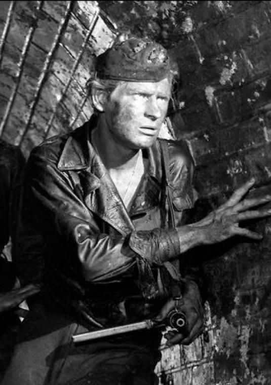 Czarno-biały kadr z filmu „Kanał” Andrzeja Wajdy przedstawiający trzech brudnych, wyczerpanych powstańców w wojskowych furażerkach, skradających się w wąskim, ceglanym kanale ściekowym.